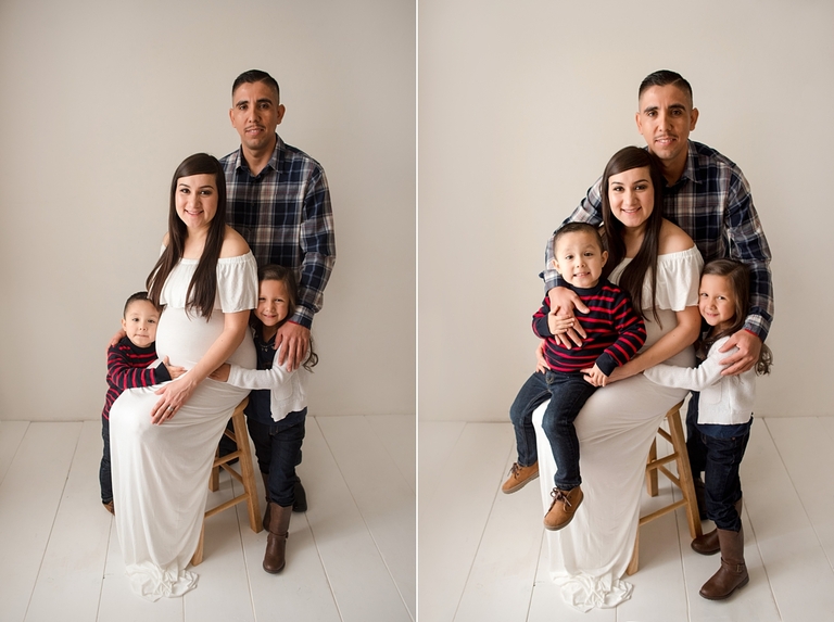 Family maternity pose with husband and children, on stool. Mom wearing white maternity gown. Posed indoor in white studio setting.